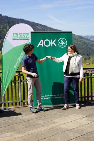 Zwei Menschen geben sich die Faust. Sie stehen aus der Terrasse der Jugendbildungsstätte in Bad Hindelang vor jeweils einem Werberaufsteller der AOK und der JDAV. Weiter hinter ihnen sind die Bad Hindelanger Berge zu sehen.