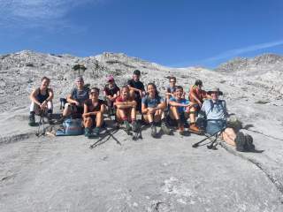 Gruppe von jungen Menschen in Wanderkleidung sitzt auf einem Felsen vor einer schroffen Felslandschaft unter blauem Himmel.
