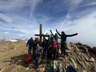 Gruppe junger Bergsteiger*innen steht um Gipfelkreuz gruppiert und reckt Hände und Eispickel in die Höhe.