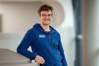 Junger Mann mit Brille und lockigem Haar lehnt lächelnd an einem modernen Geländer und trägt eine blaue Jacke mit DAV-Logo.