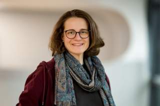 Porträt einer lächelnden Frau mit kurzen braunen Haaren, Brille, einem gemusterten Schal und einem burgunderfarbenen Pullover vor einem unscharfen hellen Hintergrund.