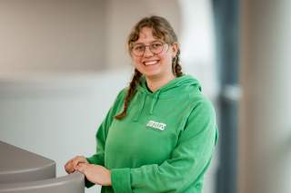 Porträt einer lächelnden Person mit Zöpfen, Brille und grünem Hoodie, die sich an einer grauen Oberfläche abstützt.