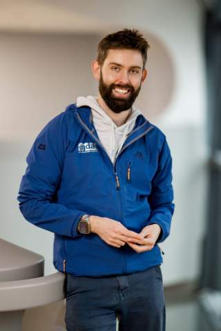 Porträt einer lächelnden Person mit Bart, die eine blaue Jacke und eine graue Kapuzenjacke trägt und entspannt an einem Geländer steht.