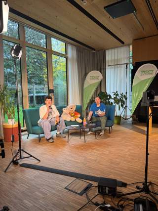 Zwei Personen sitzen in einem modern eingerichteten Raum auf blauen Sofas, hinter ihnen große Fenster und grüne Pflanzen. Auf dem Tisch stehen Getränke, daneben liegen Stofftiere. Im Hintergrund stehen zwei grüne Banner mit der Aufschrift „jdav“. Studiobeleuchtung und Kameraequipment sind sichtbar.