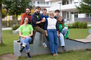 Sieben junge Menschen sitzen nebeneinander auf einer Beton-/Holz-Sitzkonstruktion auf einer Grünfläche vor einem Gebäude.