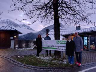 Vier Personen stehen bei einem Schild mit der Aufschrift „Jugendbildungsstätte Hindelang“ vor verschneiten Bergen und Gebäuden. Ein kahler Baum ragt in die Bildmitte.