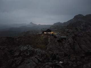 Berghütte mit beleuchteten Fenstern in einer grauen Berglandschaft und Wolken, es ist ziemlich dunkel.