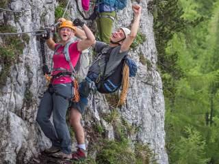 Zwei Personen am Klettersteig, eine Hand am Seil, die andere als Faust in die Luft gereckt