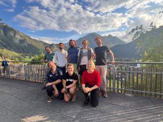 Act junge Menschen stehen in zwei Reihen vor einem Geländer. Hinter dem Geländer befindet sich eine Landschaft mit Bergen, der Himmel ist bewölkt.