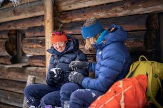 Zwei Menschen sitzen im Winter vor einer Hütte mit Daunenkleidung von Mountain Equipment.