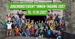 Gruppenfoto von etwa 50 jungen Menschen vor einer alten Steinmauer, alle heben die Arme; darüber ein grünes Banner mit der Aufschrift 