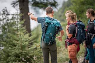 4 Personen mit Rucksäcken und Wanderkleidung stehen im Wald und gucken in eine Richtung, eine Person zeigt in diese Richtung.