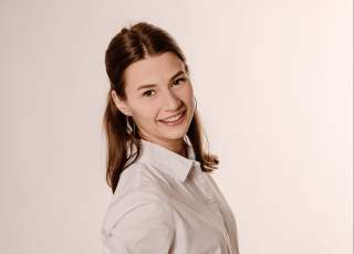 Junge Frau mit braunen Haaren, weißem Hemd und blauer Jeans steht lächelnd vor hellem Hintergrund.