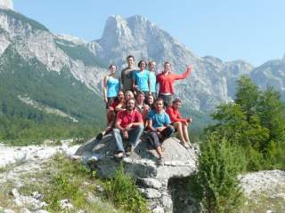 Eine Gruppe junger Erwachsener sitzt auf einem großen Stein in den Bergen; im Hintergund eine Bergkette