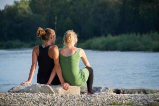 Zwei Frauen sitzen eng aneinander auf einem Stein am Isarstrand und schauen auf den Fluss