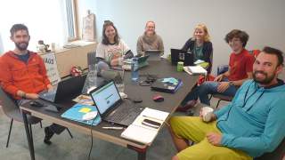 Eine Gruppe von sechs Personen sitzt an einem Tisch mit Laptops und Notizbüchern