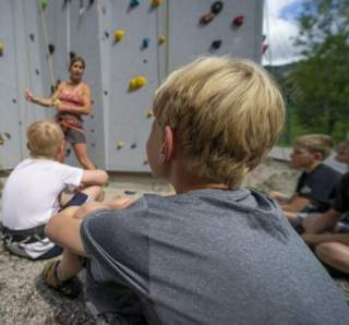 Kinder sitzen aufmerksam vor einer Kletterwand und hören einer Trainerin zu, die Kletteranweisungen gibt.