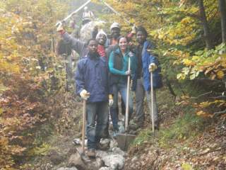 Eine diverse Gruppe aus Menschen steht auf einem Bergweg. Sie halten Werkzeuge in der Hand