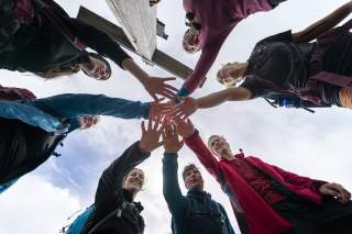 Menschen stehen im Kreis neben einem Gipfelkreuz und strecken je eine Hand in die Mitte des Kreises. Das Bild ist von untern aufgenommen.