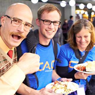 Drei Personen sitzen gemeinsam an einem Tisch und lachen, zwei von ihnen tragen blaue T-Shirts und halten Teller mit Essen.