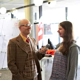 Ein Mann mit Brille und kariertem Sakko hält ein Mikrofon und spricht mit einer lächelnden jungen Frau mit langem Haar in einem hellen Innenraum.