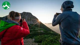 Zwei Wander*innen machen Fotos von der Sonne, die hinter einem Berg hervorkommt