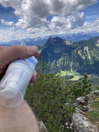 Ein Wanderer hält Sonnencreme in der Hand, im Hintergrund eine malerische Berglandschaft mit grünen Wäldern und schneebedeckten Gipfeln.