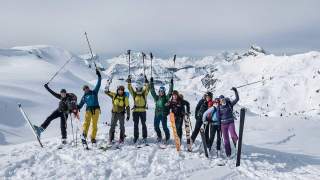 Eine Gruppe von Skitourengehern steht jubelnd auf einem verschneiten Berggipfel in den Alpen, umgeben von einer beeindruckenden winterlichen Berglandschaft.