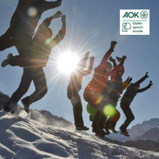 Eine Gruppe von Menschen springt im Schnee auf einem Berggipfel in die Luft, die Sonne scheint im Hintergrund