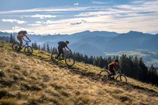 Drei Jugendliche fahren mit dem Mountainbike eine Bergwiese runter; im Hintergrund eine Berglandschaft