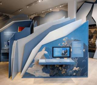 Ein modernes Ausstellungsdisplay mit blauen, geometrischen Strukturen und einem Bildschirm, das Informationen zur Gemeinschaft und Naturschutz in deutscher und englischer Sprache präsentiert.
