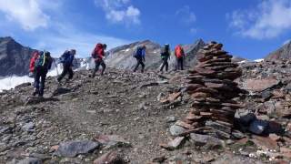 Eine Gruppe von Menschen wandert durch eine felsige Landschaft. Im Hintergrund sind Berggipfel zu sehen. Im Vordergrund sieht man einen Steinhaufen