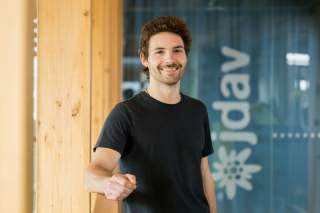 Mann mit kurzen braunen Locken und Bart steht lächelnd an einem Holzgeländer