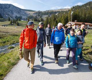 Mehrere Menschen wandern nebeneinander auf breitem Fahrweg, im Hintergrund eine Hütte, neben dem Weg ein Bach.