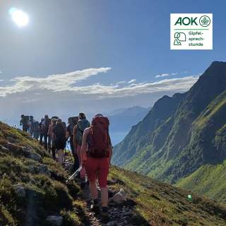 Eine Gruppe Menschen wandert hintereinander einen Bergpfad entlang. Alle haben Rucksäcke auf dem Rücken.